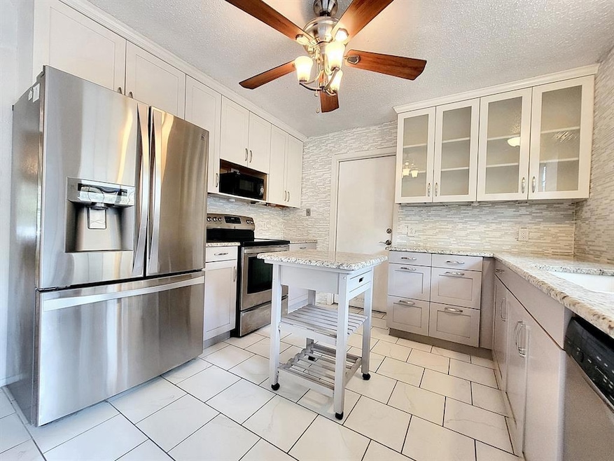 Kitchen with stainless steel appliances, a textured ceiling, glass insert cabinets, light stone countertops, and decorative backsplash