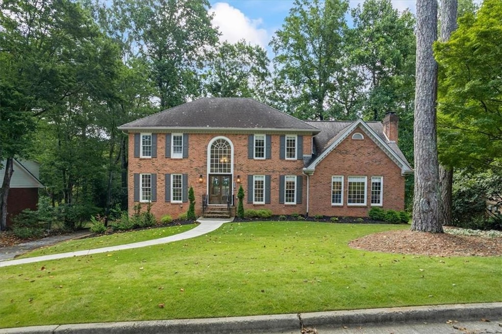 Colonial-style house with a front yard, brick siding, a chimney, and view of wooded area