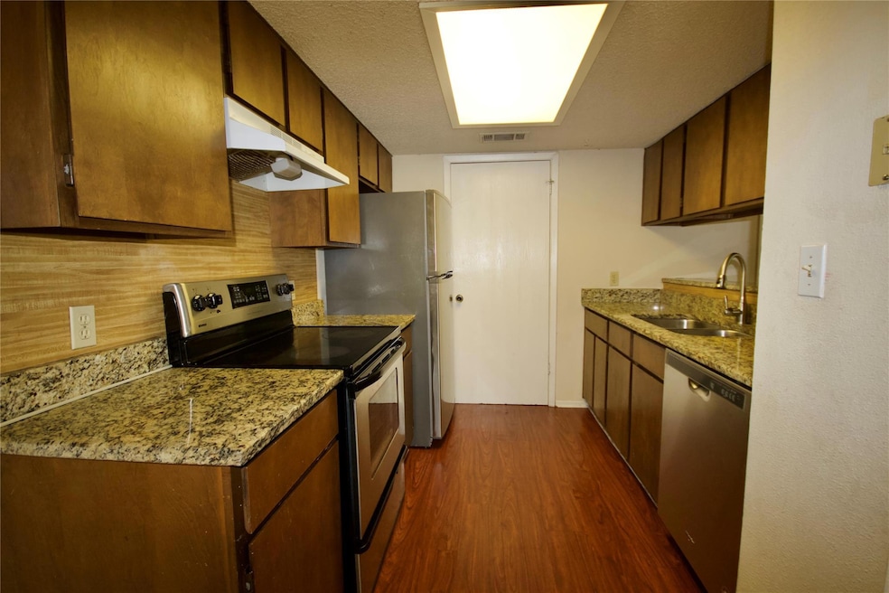 Kitchen featuring appliances with stainless steel finishes, dark wood-style floors, light stone counters, under cabinet range hood, and brown cabinets