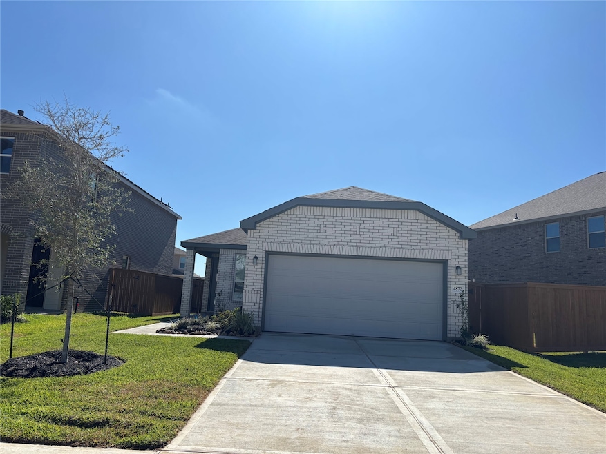 This photo shows a modern, single-story brick house with a two-car garage. The home features a well-maintained front yard with a small tree and neat landscaping. It's set in a neighborhood with similar homes and has a clear, sunny sky overhead.