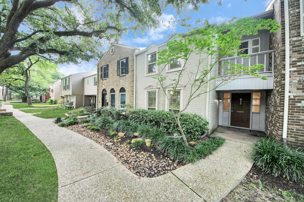 This charming townhouse features a welcoming stone and brick exterior with lush landscaping. A pathway leads to the front entrance, shaded by mature trees, creating a serene and inviting atmosphere.