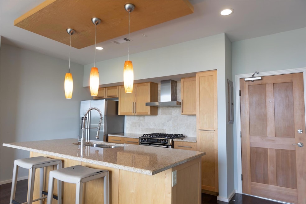Kitchen featuring light brown cabinets, hanging light fixtures, tasteful backsplash, wall chimney range hood, and a kitchen island with sink