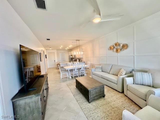 Living room with visible vents, vaulted ceiling, ceiling fan with notable chandelier, light tile patterned flooring, and a decorative wall