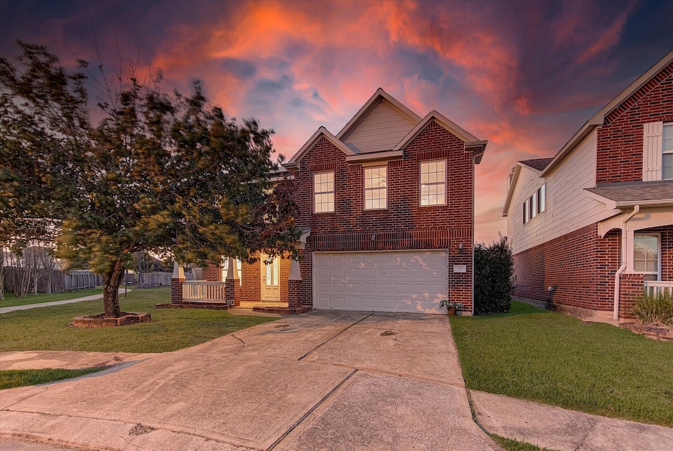 This photo showcases a two-story brick home with a double garage and a cozy front porch. The driveway is spacious, and a well-maintained lawn surrounds the property. The sky is beautifully lit with vibrant sunset colors, enhancing the home's curb appeal.
