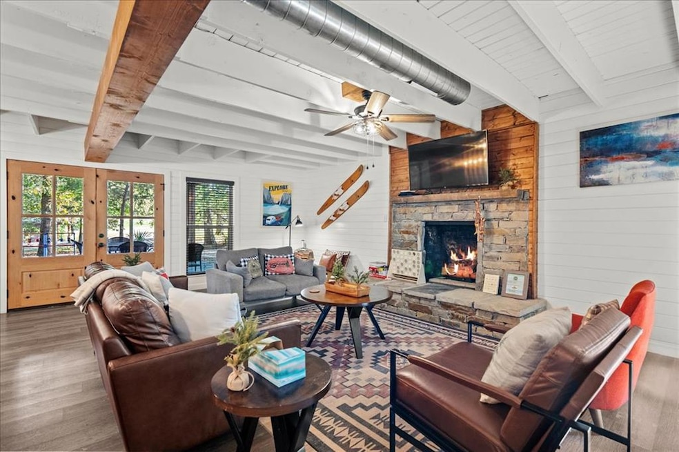 Living area with beamed ceiling, wooden walls, wood finished floors, a ceiling fan, and a stone fireplace