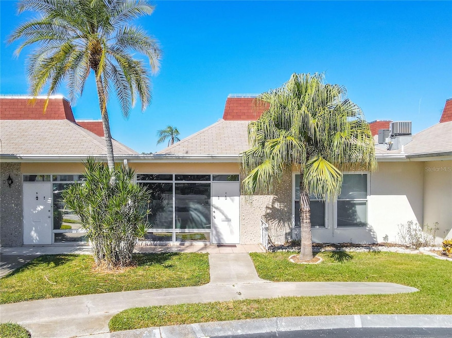 A welcoming condo entrance features a neatly paved sidewalk lined with tall, graceful palm trees on either side, creating a tropical and inviting approach. The pathway leads up to a sleek storm door that offers both style and protection, accented by a transom window above that allows natural light to filter into the entryway. The combination of greenery and clean architectural details gives the entrance a bright, resort-like charm while maintaining a sense of island elegance.