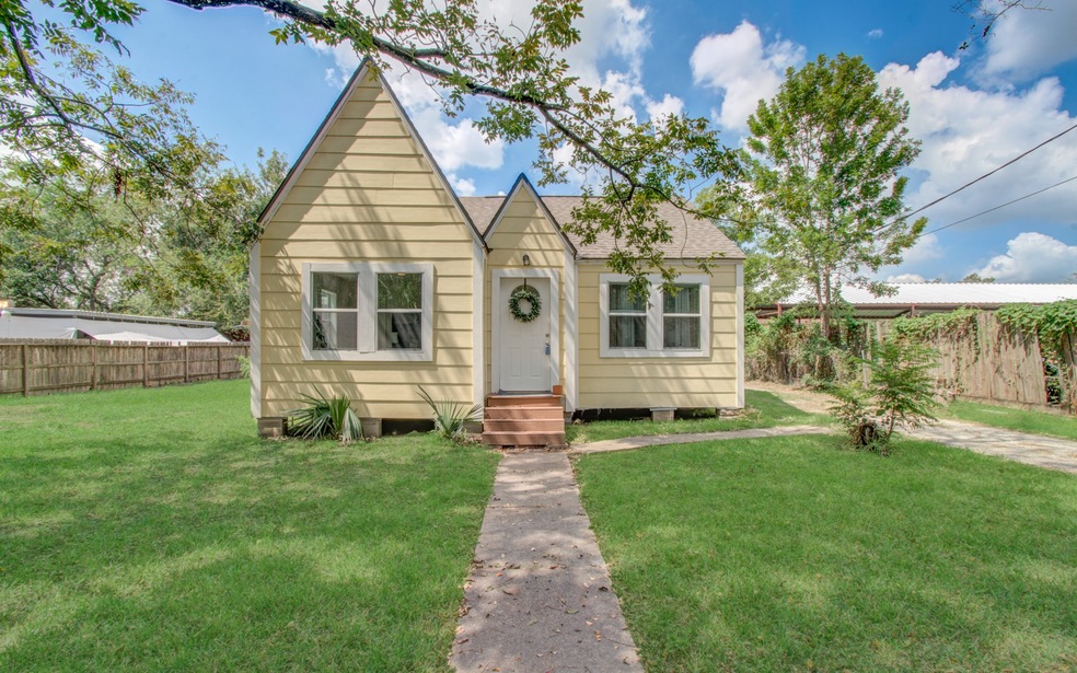 Charming single-story home featuring a peaked roof, light yellow siding, and white trim. The property includes a welcoming front door with a decorative wreath, a well-maintained lawn, and a clear walkway leading up to the house. A wooden privacy fence surrounds the spacious backyard.