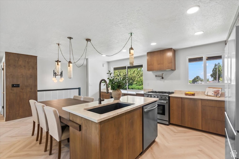 Kitchen with brown cabinets, an island with sink, pendant lighting, stainless steel appliances, and a textured ceiling