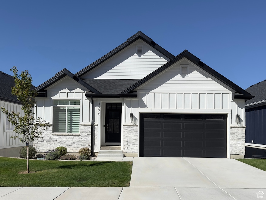 Modern farmhouse style home with board and batten siding, a garage, driveway, stone siding, and a front lawn