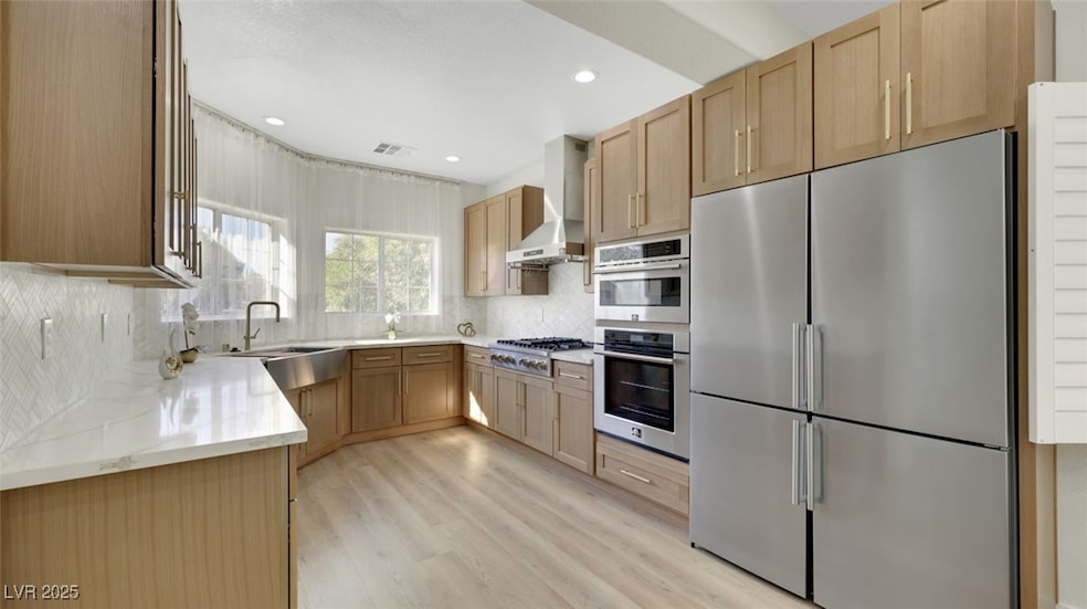 Kitchen with stainless steel appliances, light brown cabinetry, wall chimney range hood, tasteful backsplash, and light stone countertops