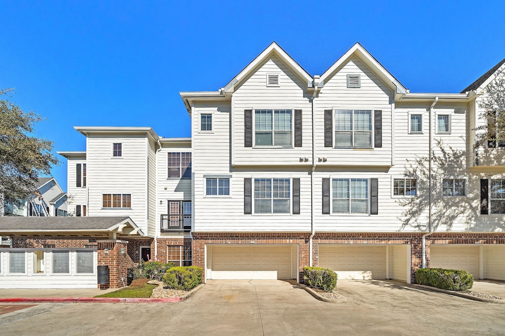 This photo shows a modern townhouse/condominium complex with crisp white siding and brick accents. The building features multiple stories, large windows for natural light, and attached garages. The driveway is spacious, and there are small landscaped areas with shrubs.