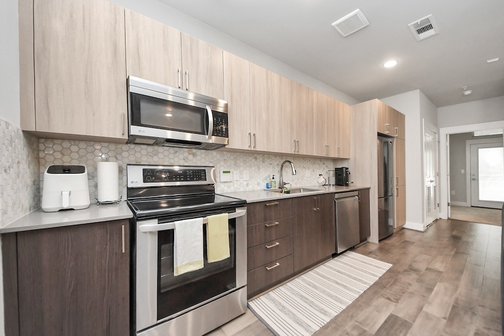 This kitchen features modern cabinetry with light wood tones, stainless steel appliances, and a sleek backsplash. The open layout provides ample counter space and a streamlined appearance.