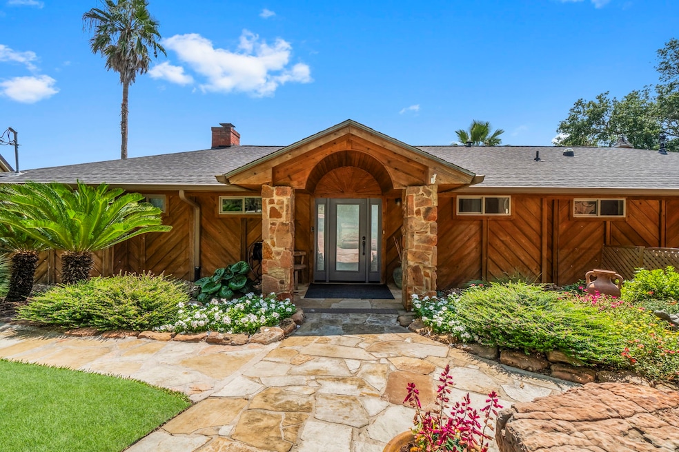 Charming single-story home with a rustic stone and wood exterior. Features a welcoming entrance, lush landscaping, and a paved walkway surrounded by vibrant flowers and greenery. Perfect for nature lovers seeking a cozy and inviting atmosphere.