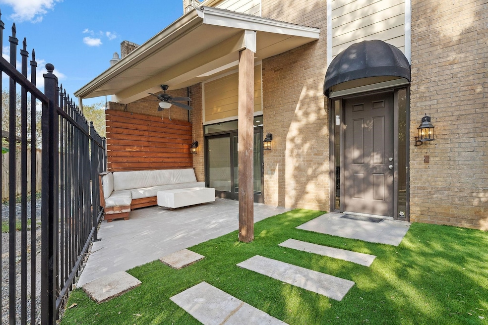 This photo showcases a cozy outdoor patio with a partially covered seating area featuring a modern L-shaped couch. It has a wooden privacy panel, ceiling fan, and is adjacent to a brick wall with a stylish entrance. A small, well-maintained lawn with stepping stones leads to the front door, all enclosed by a wrought iron fence.