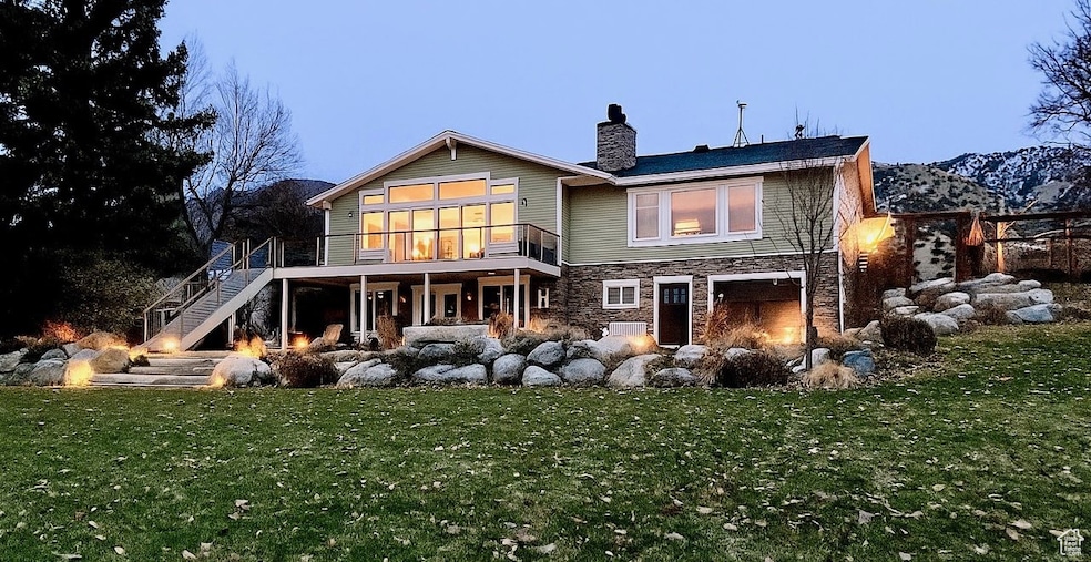 Back of house at dusk featuring stairs, stone siding, a chimney, a yard, and a wooden deck