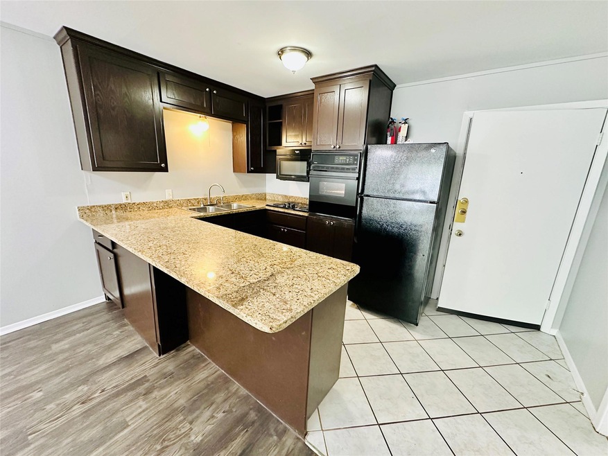 Kitchen featuring a peninsula, black appliances, dark brown cabinetry, a kitchen bar, and light stone counters