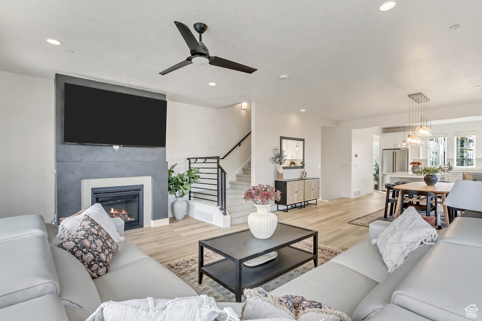 Living area featuring light wood-type flooring, a glass covered fireplace, stairs, a ceiling fan, and recessed lighting