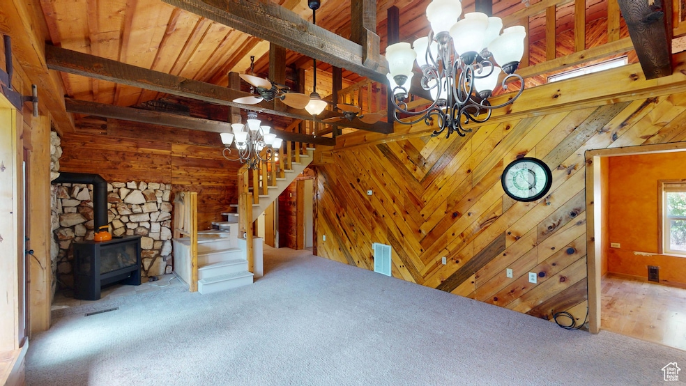 Unfurnished living room featuring a wood stove, stairway, a chandelier, carpet, and beam ceiling