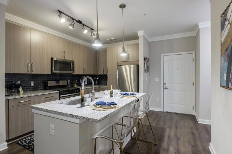 Kitchen with rail lighting, light stone counters, a kitchen island with sink, stainless steel appliances, and sink