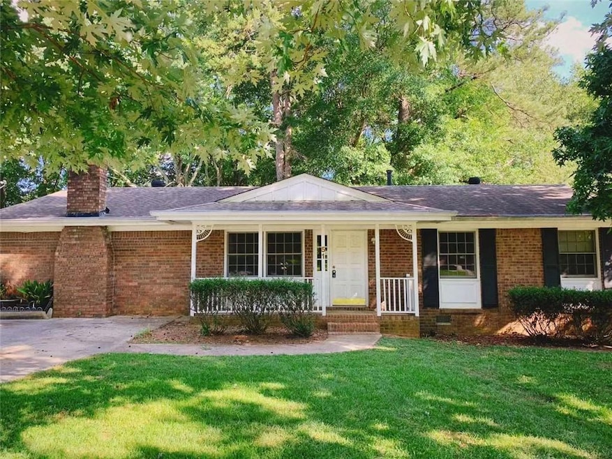 Single story home with a chimney, brick siding, a front lawn, covered porch, and roof with shingles