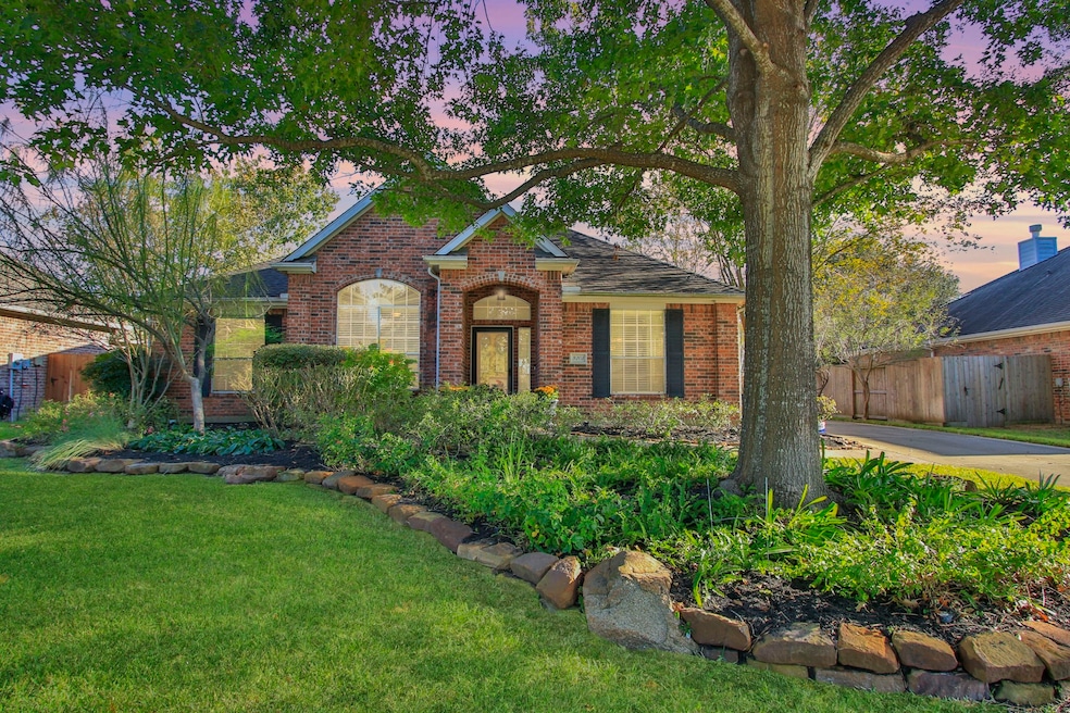 Inviting front elevation framed by mature shade trees and lush landscaping! This charming red brick one-story features classic curb appeal with arched windows, black shutters, and a welcoming covered entry.