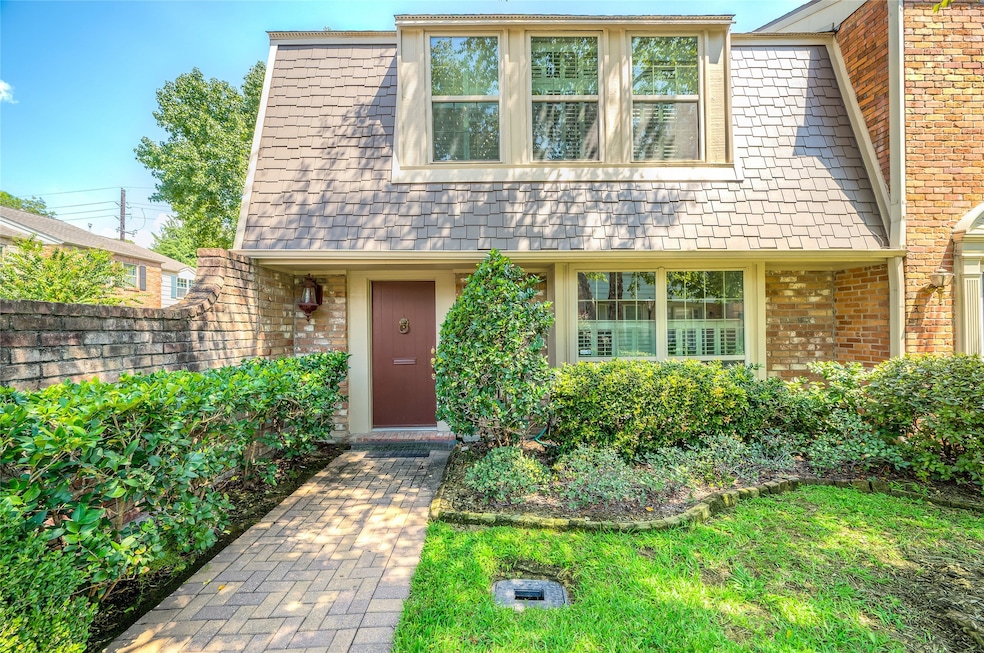 This charming two-story home features a classic brick exterior with a unique shingled roof. The front entrance is framed by lush greenery and a paved walkway, offering a welcoming and quaint curb appeal. Large windows provide ample natural light, accentuating the cozy atmosphere.