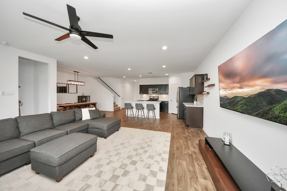 This stunning open-concept space effortlessly combines style and functionality. The modern kitchen boasts rich dark cabinetry, a large center island, and gleaming stainless steel appliances—ideal for both everyday meals and entertaining. It flows seamlessly into the spacious living area, featuring wood flooring, a comfortable sectional sofa, and a wall-mounted TV. A nearby dining area and an elegant staircase add to the thoughtful layout, all enhanced by sleek lighting and a contemporary ceiling fan that co