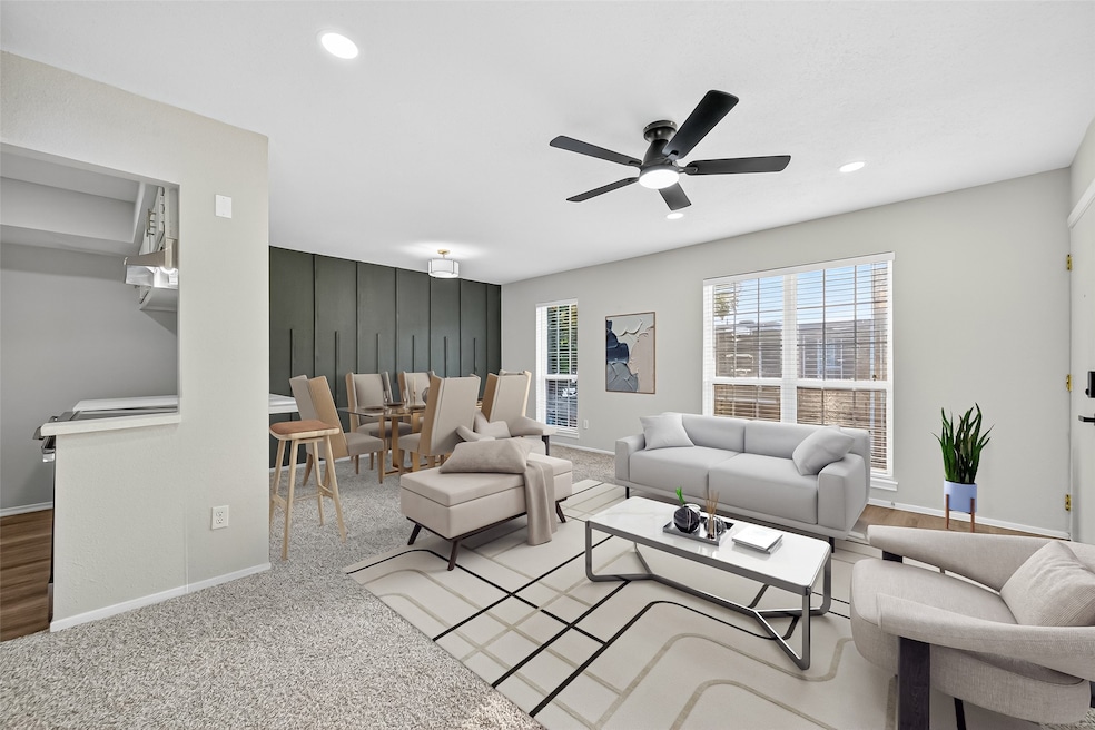 This photo showcases a modern, open-plan living and dining area with neutral tones. It features a comfortable seating arrangement, a ceiling fan, and large windows that let in plenty of natural light. The dining area is adjacent to the kitchen, which has a small breakfast bar. The space is stylishly furnished with contemporary decor.