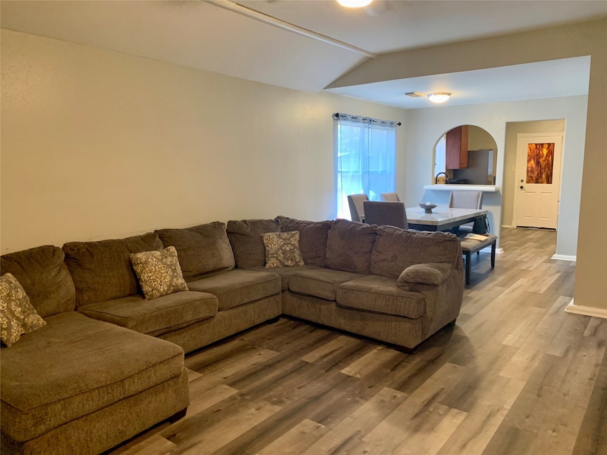 View of living/dining open concept room with vaulted ceiling and natural light. View through kitchen to back door entrance to the back covered patio. Recently installed flooring.