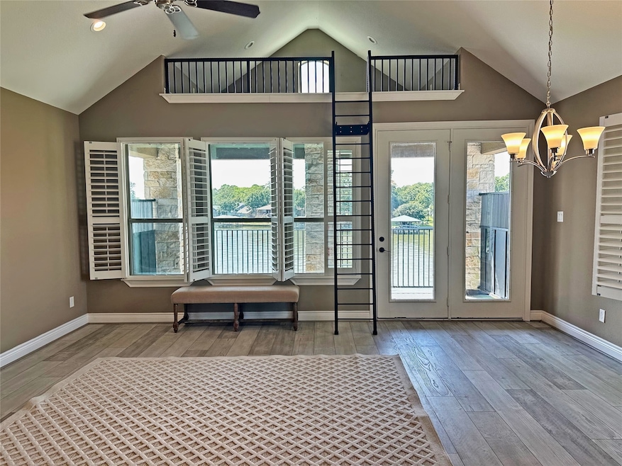 This room features high ceilings with a ceiling fan, large windows with shutters offering a scenic water view, and a loft area accessible by a wall ladder. The space is bright and inviting, with wood flooring and a modern chandelier.