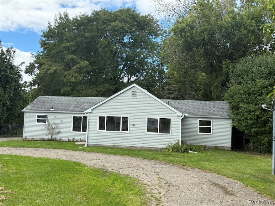 Single story home with a shingled roof, a front lawn, driveway, and view of wooded area
