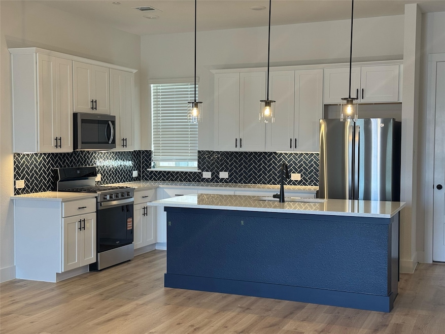 Kitchen with white cabinetry, stainless steel appliances, hanging light fixtures, a center island with sink, and light stone counters
