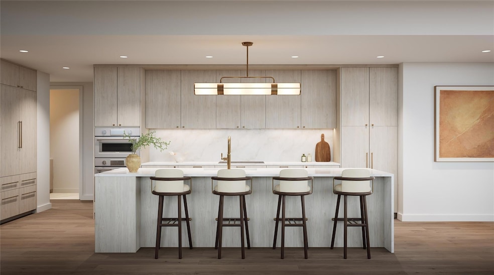 Kitchen in Tranquil palette featuring a large breakfast bar, designer lighting, wood flooring, recessed lights, and natural stone quartzite countertops. Fully integrated Thermador appliance suite — including refrigerator, induction cooktop, wall oven, dishwasher, and built-in wine preservation tower — all standard, not upgrade. Soft, light-toned cabinetry and subtle detailing create an airy, refined aesthetic. Representative finish shown; three palettes available, design varies by residence.
