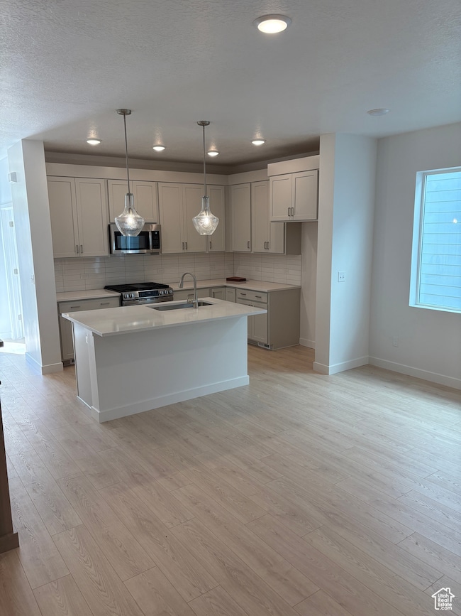 Kitchen with backsplash, pendant lighting, a kitchen island with sink, stainless steel appliances, and light wood-style flooring