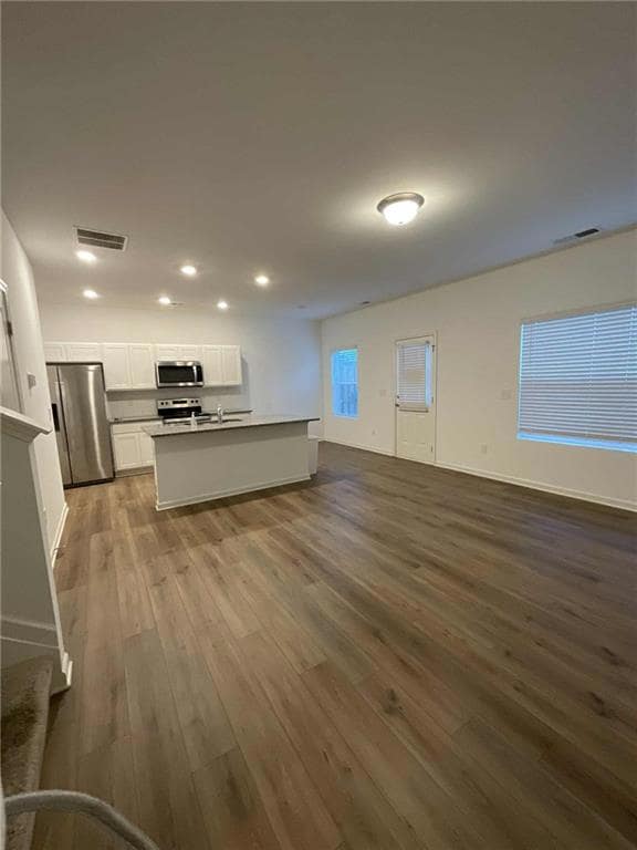 Kitchen featuring white cabinets, stainless steel appliances, open floor plan, a center island with sink, and dark wood-style flooring