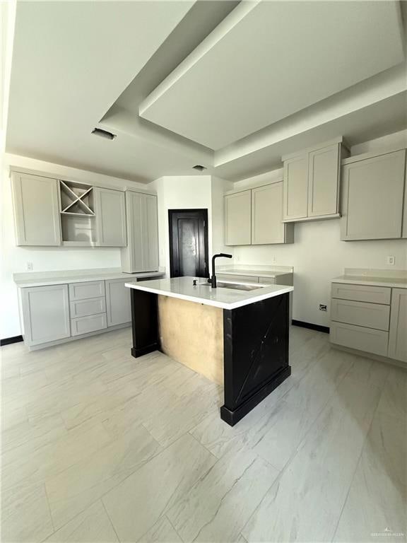 Kitchen with a sink, gray cabinetry, a raised ceiling, light marble finish flooring, and an island with sink