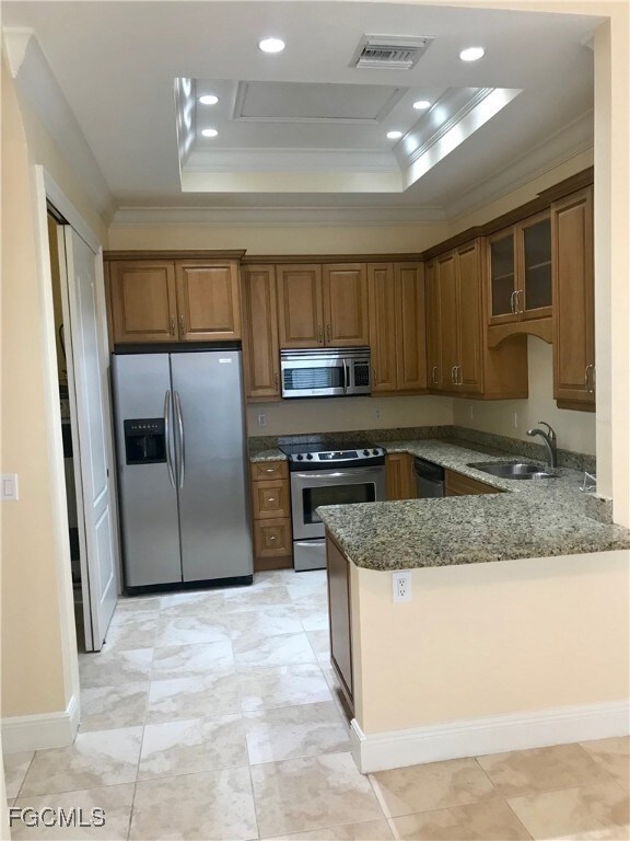 Kitchen featuring a raised ceiling, ornamental molding, brown cabinetry, dark stone counters, and appliances with stainless steel finishes