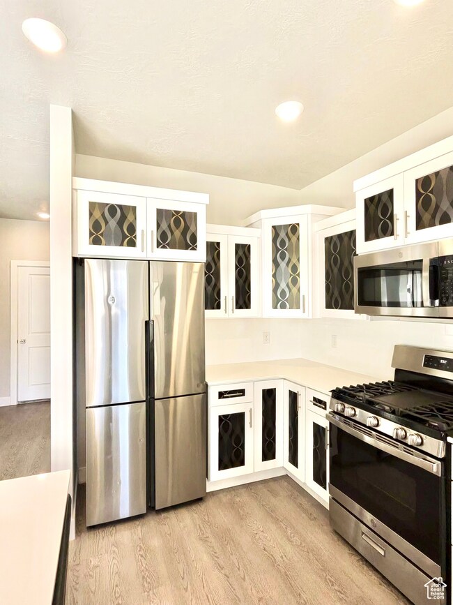 Kitchen featuring appliances with stainless steel finishes, light countertops, white cabinetry, glass insert cabinets, and light wood finished floors