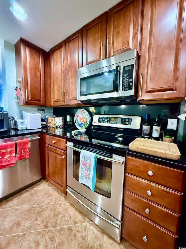 Kitchen featuring appliances with stainless steel finishes, brown cabinets, light tile patterned floors, and dark countertops