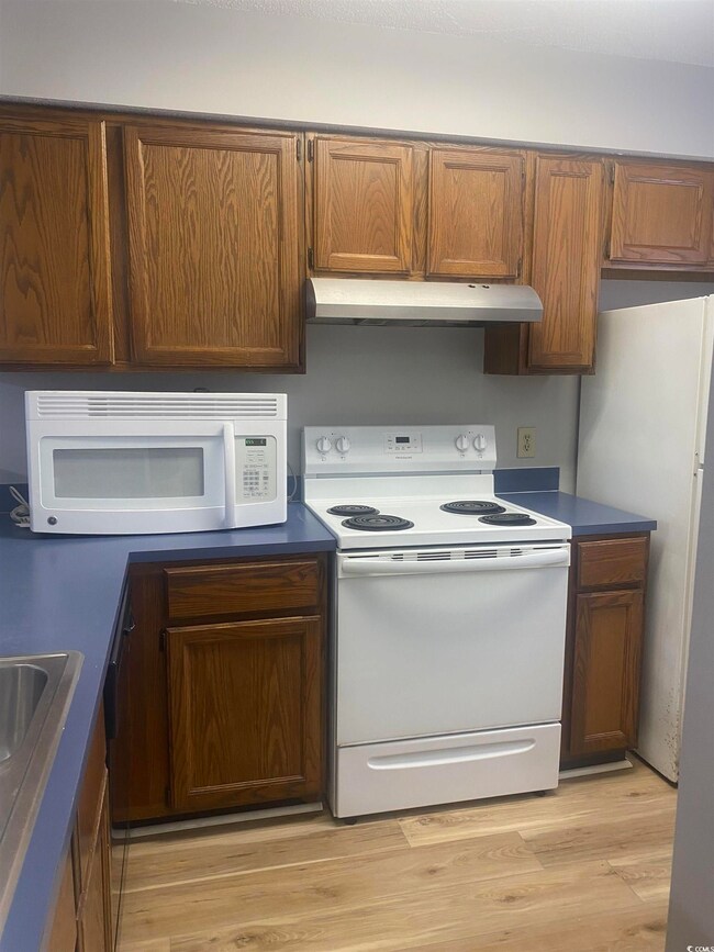 Kitchen with white appliances, under cabinet range hood, light wood-style flooring, brown cabinets, and dark countertops