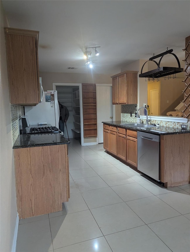 Kitchen with decorative backsplash, brown cabinetry, dark stone counters, stainless steel dishwasher, and light tile patterned floors