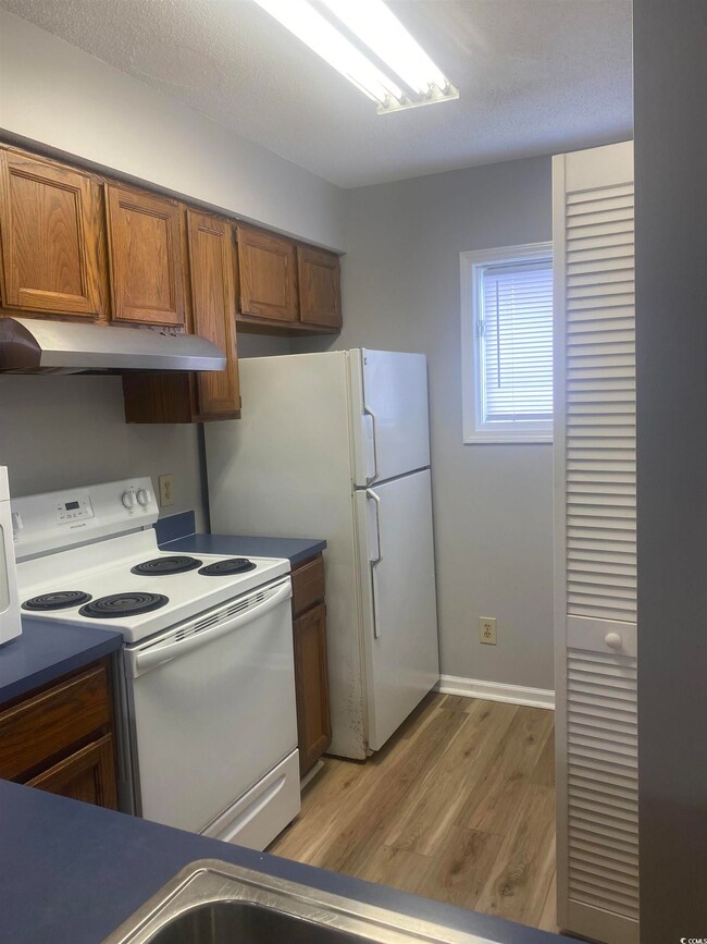Kitchen with white electric stove, under cabinet range hood, light wood-style flooring, brown cabinets, and dark countertops