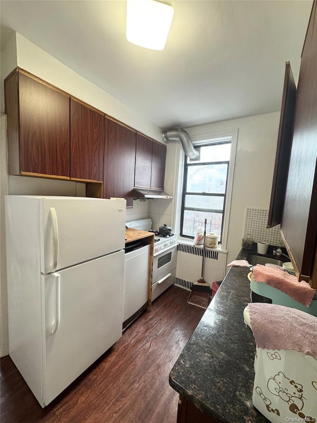 Kitchen featuring white appliances, radiator, dark wood-type flooring, dark brown cabinets, and under cabinet range hood