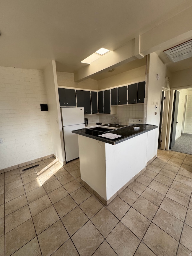 Kitchen featuring dark cabinetry, backsplash, freestanding refrigerator, a peninsula, and light tile patterned flooring