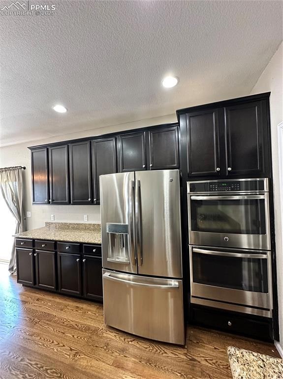 Kitchen featuring stainless steel refrigerator with ice dispenser, double wall oven, dark cabinetry, a textured ceiling, and recessed lighting