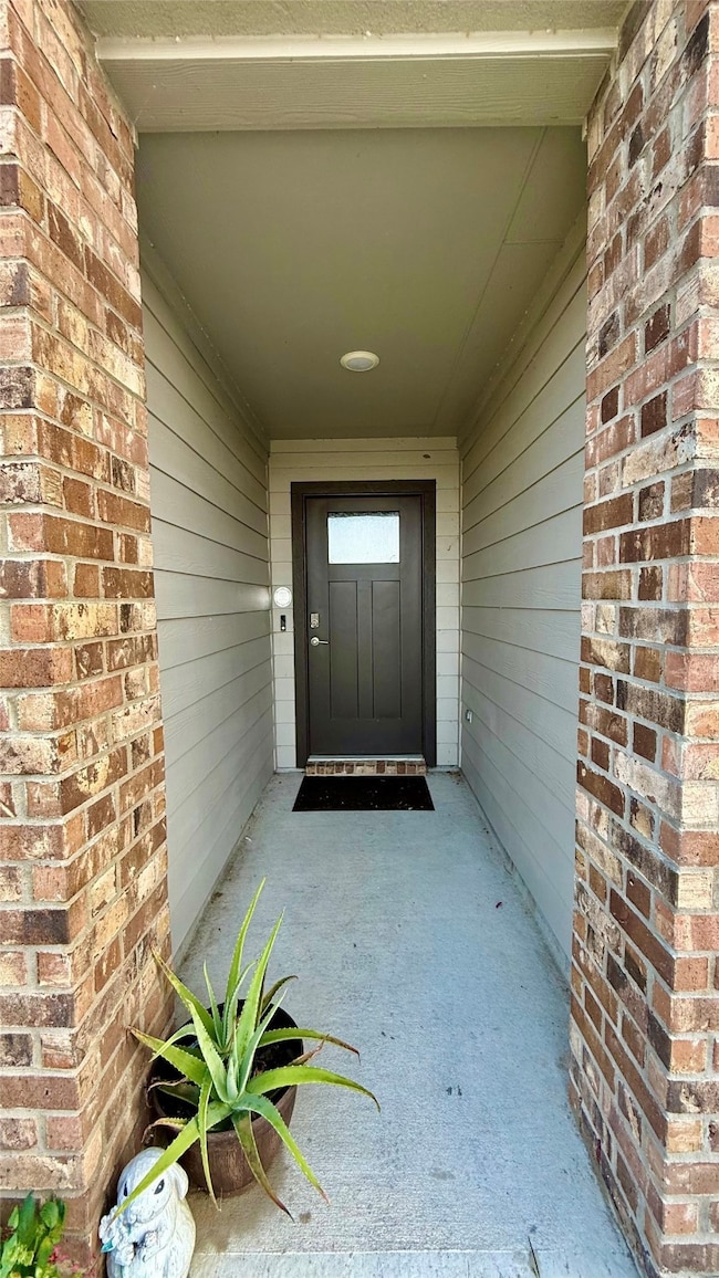 This inviting entryway features a sheltered porch with brick columns and siding, leading to a modern front door with a frosted glass panel. The space is accented by a potted plant, adding a touch of greenery.