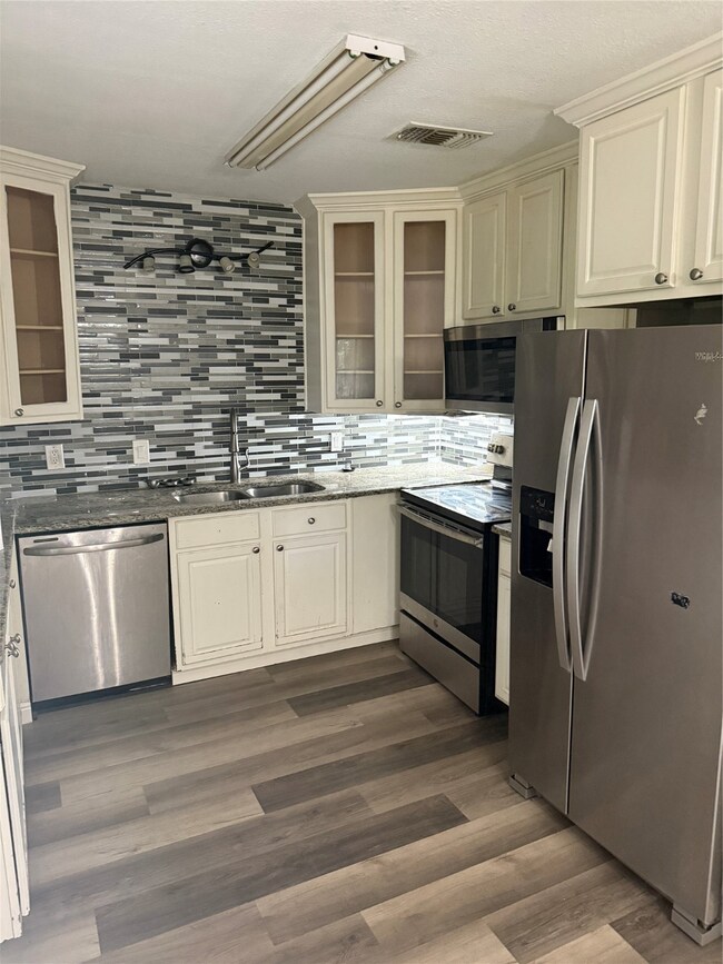 Kitchen featuring appliances with stainless steel finishes, glass insert cabinets, tasteful backsplash, dark stone counters, and dark wood-style flooring