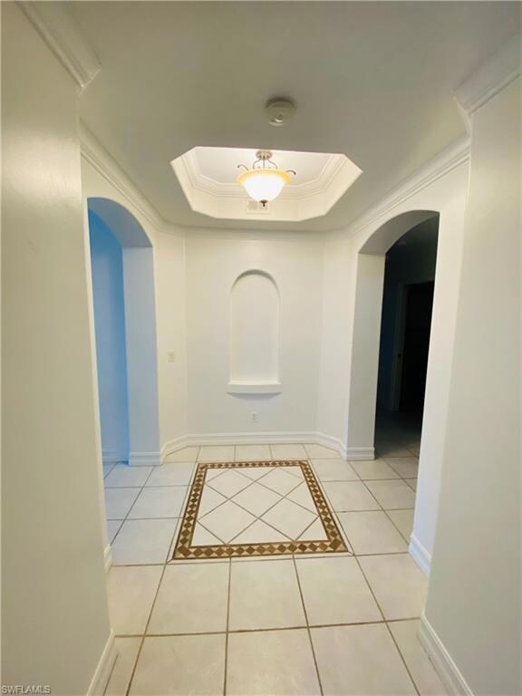 Hallway featuring arched walkways, light tile patterned floors, a raised ceiling, crown molding, and inlaid floor details
