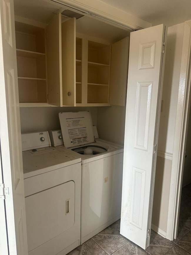 Compact laundry area with a washer and dryer set, enclosed by white bifold doors. Above the appliances are open cabinets for storage. The space features patterned tile flooring.