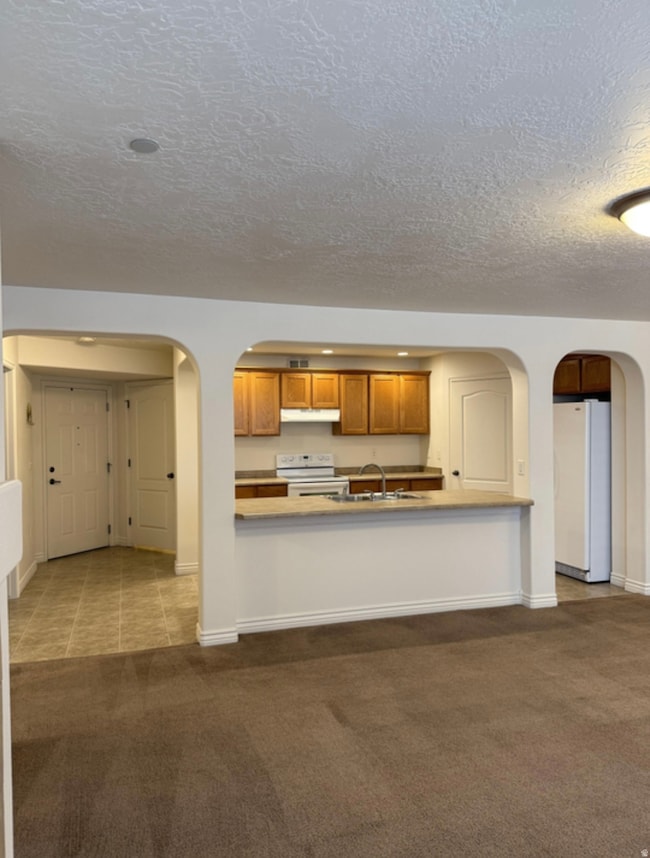 Kitchen featuring arched walkways, dark carpet, brown cabinetry, white appliances, and a textured ceiling